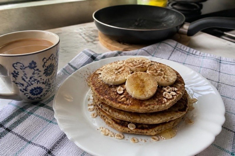 Pilha de panquecas de banana com aveia, com rodelas de banana, aveia e mel, num prato branco ao lado de uma xícara de café com leite