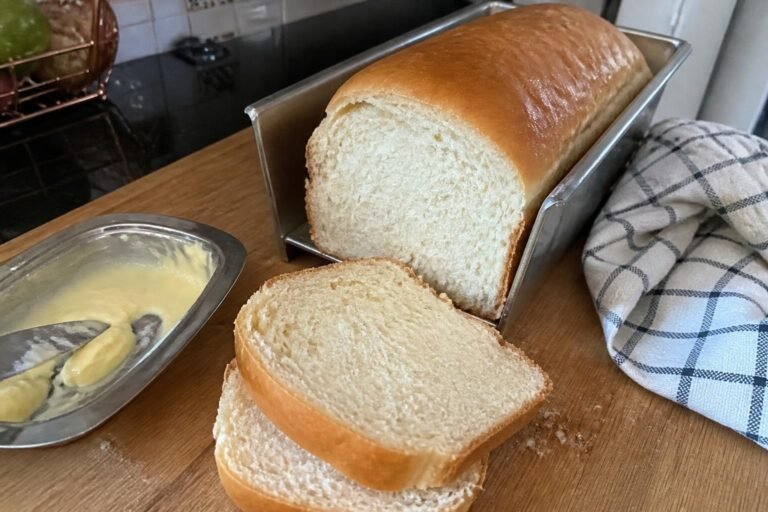 Pão caseiro de leite na forma, com duas fatias cortadas mostrando o miolo fofinho, ao lado de um pote de manteiga