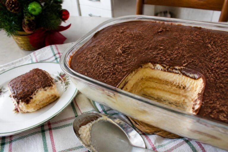 Travessa de vidro com pavê de bolacha maizena, com uma porção já servida mostrando as camadas de creme e bolacha e cobertura de chocolate